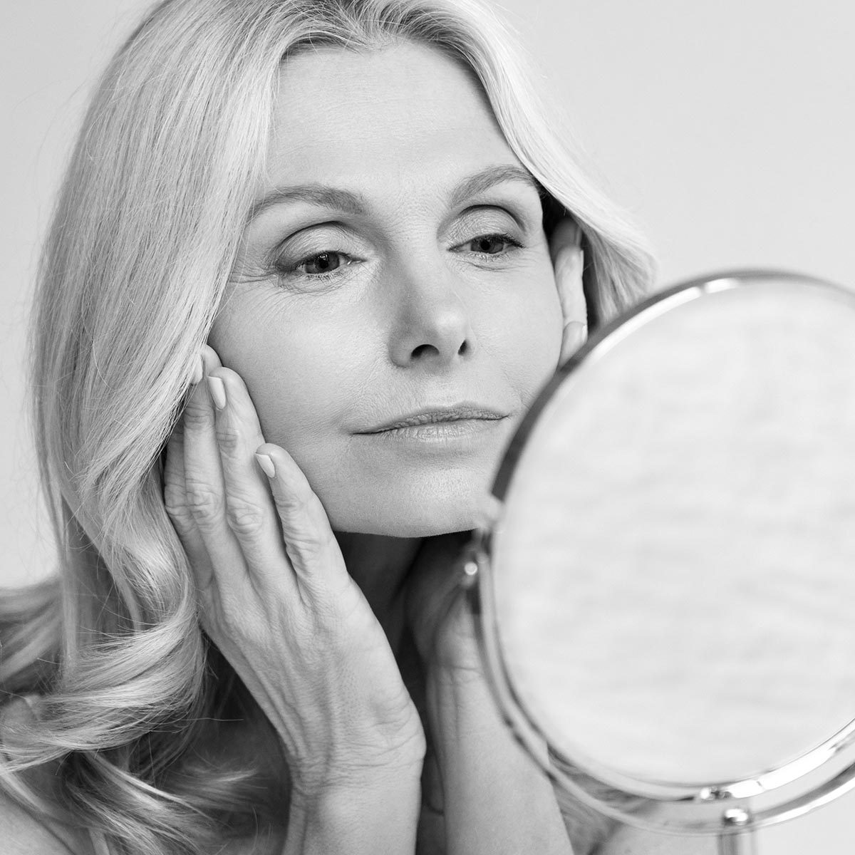 Closeup portrait of gorgeous happy middle age woman looking at mirror touching her skin enjoying treatment for dry skin. Advertising of antiaging beauty skin care products.